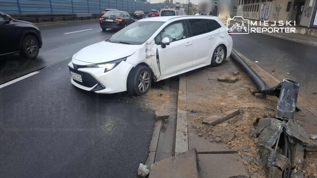 Biały samochód osobowy stoi na poboczu drogi, z uszkodzoną lewą stroną i otwartymi drzwiami. W tle widać inne pojazdy na jezdni.