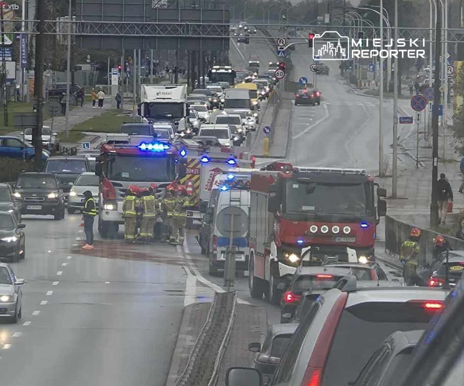 Grupa strażaków w mundurach omawia sytuację obok wozu strażackiego na ruchliwej ulicy, w tle zablokowany ruch samochodowy.