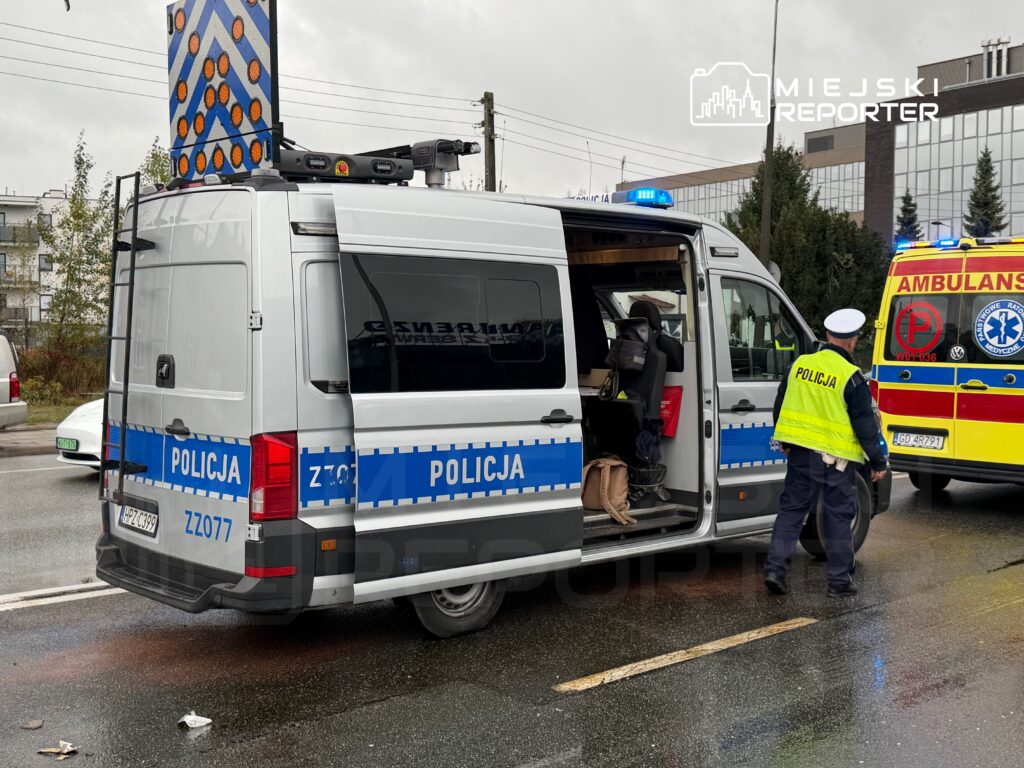 Policyjny samochód dostawczy z włączonymi sygnalizatorami, obok mundurowy kontrolujący sytuację na drodze.
