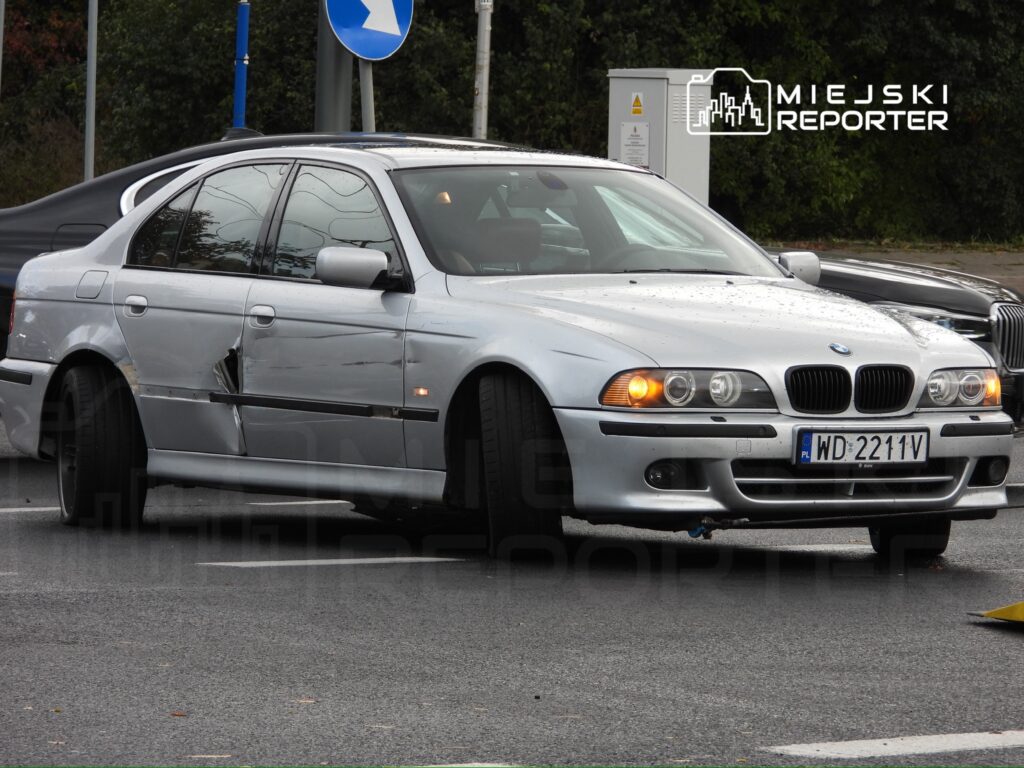 Uszkodzony srebrny samochód osobowy marki BMW stoi na skrzyżowaniu, w tle widać inne pojazdy.