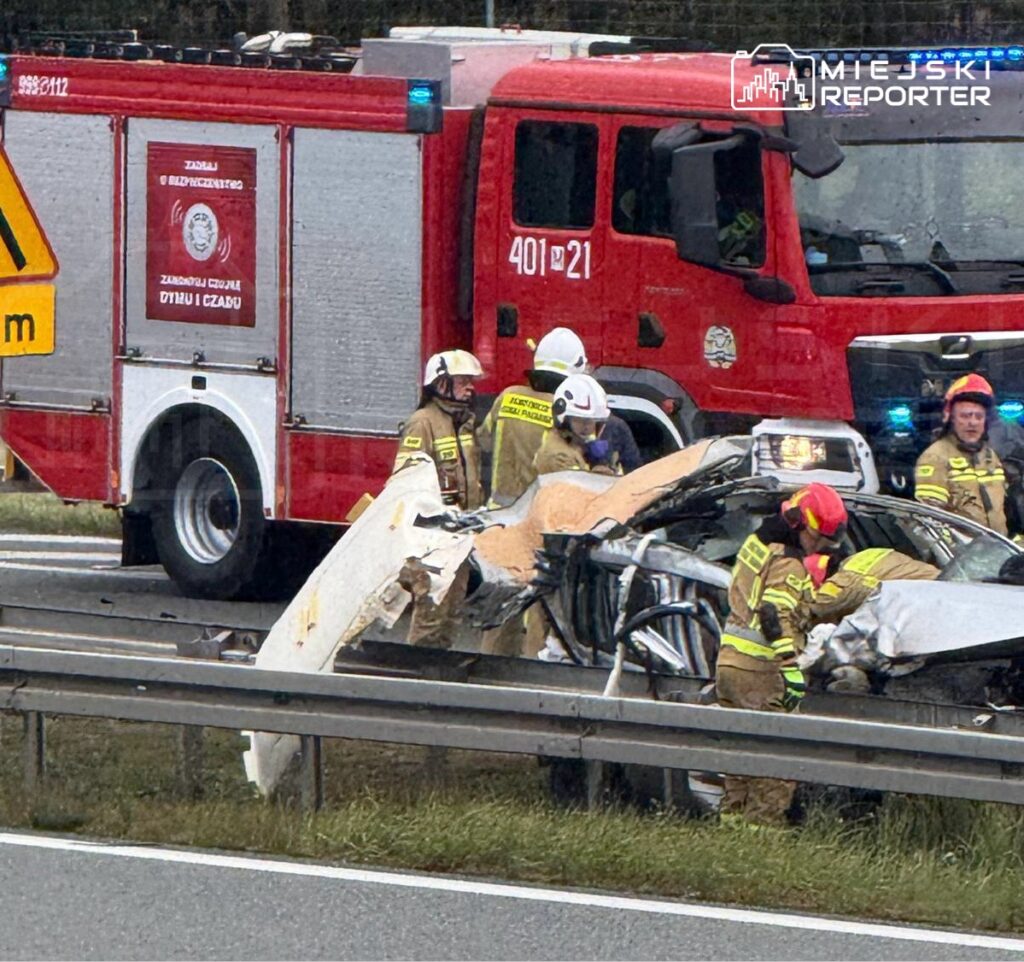 Strażacy pracują nad rozbitym samochodem osobowym przy drodze, obok stoi wóz strażacki z włączonymi sygnałami.