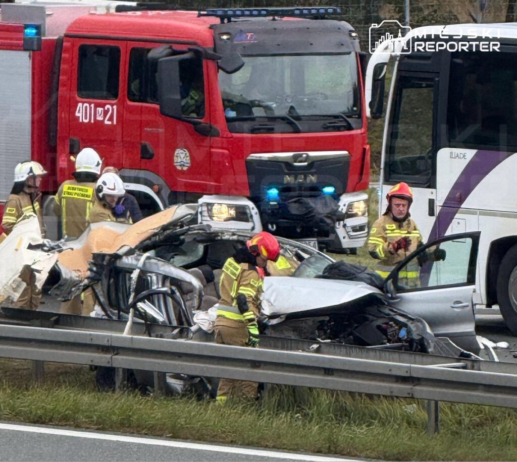 Strażacy w kaskach i mundurach pracują przy rozbitym samochodzie na autostradzie, obok widać woz strażacki.