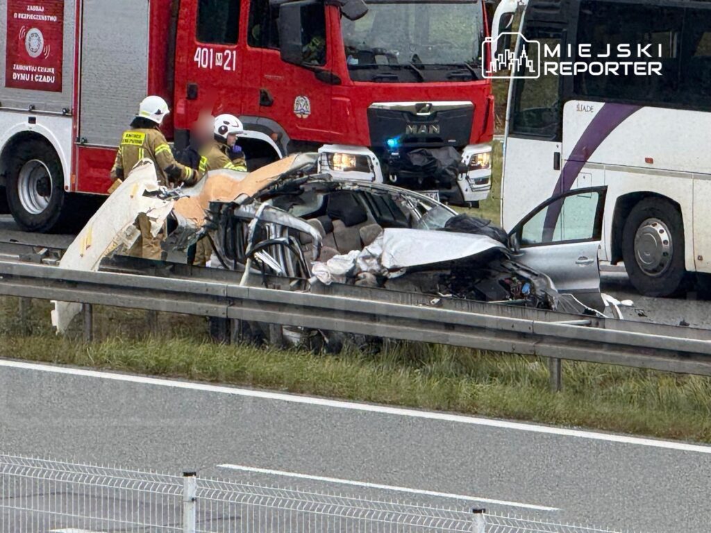 Strażacy pracują nad usunięciem zniszczonego pojazdu osobowego przy drodze, obok wozu strażackiego.