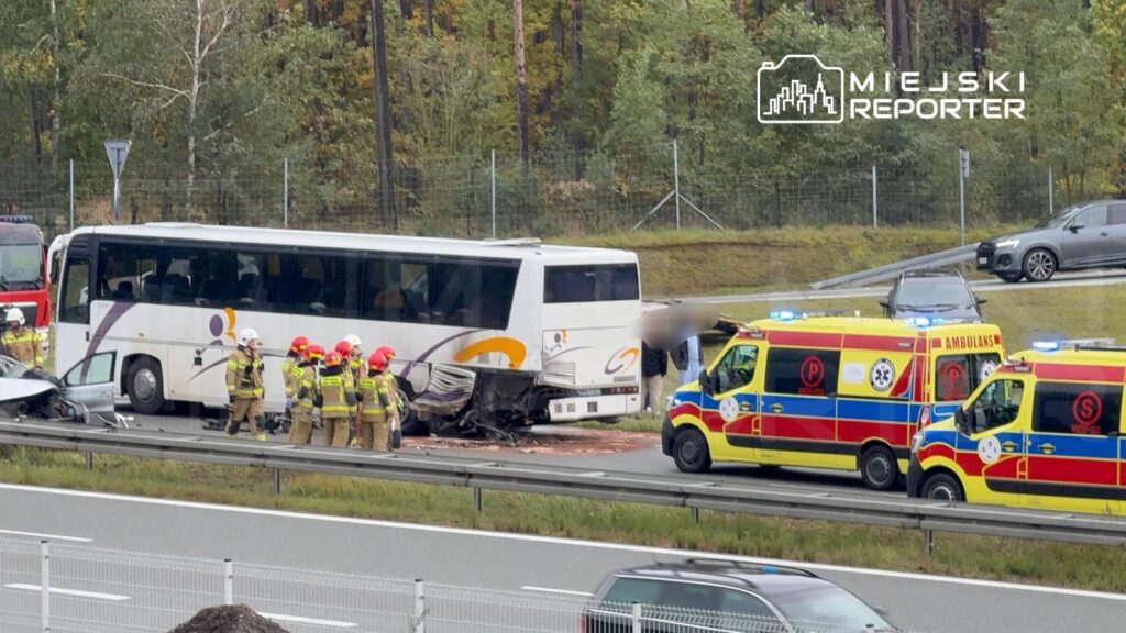 Grupa strażaków w hełmach udziela pomocy poszkodowanym przy zderzonym autobusie na drodze ekspresowej.