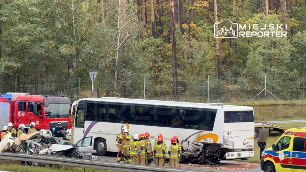 Wielu strażaków pracuje przy zderzeniu autobusu z osobowym samochodem na drodze, otoczeni przez pojazdy ratunkowe i las.