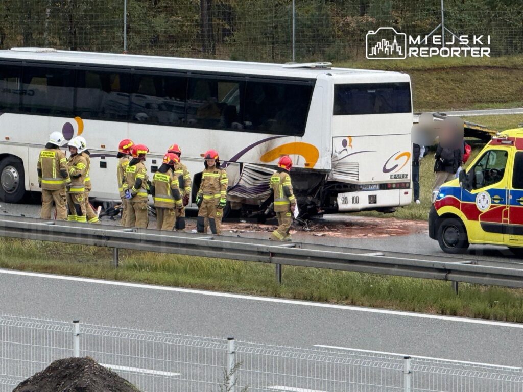 Grupa strażaków w ubraniach ochronnych prowadzi akcję ratunkową przy uszkodzonym autobusie na drodze, obok stoi karetka.