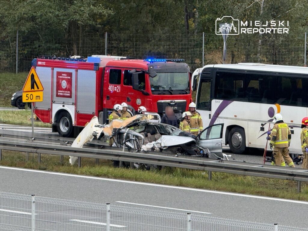 Ratownicy w kaskach pracują przy zniszczonym samochodzie po wypadku drogowym obok strażackiego wozu i autobusu.
