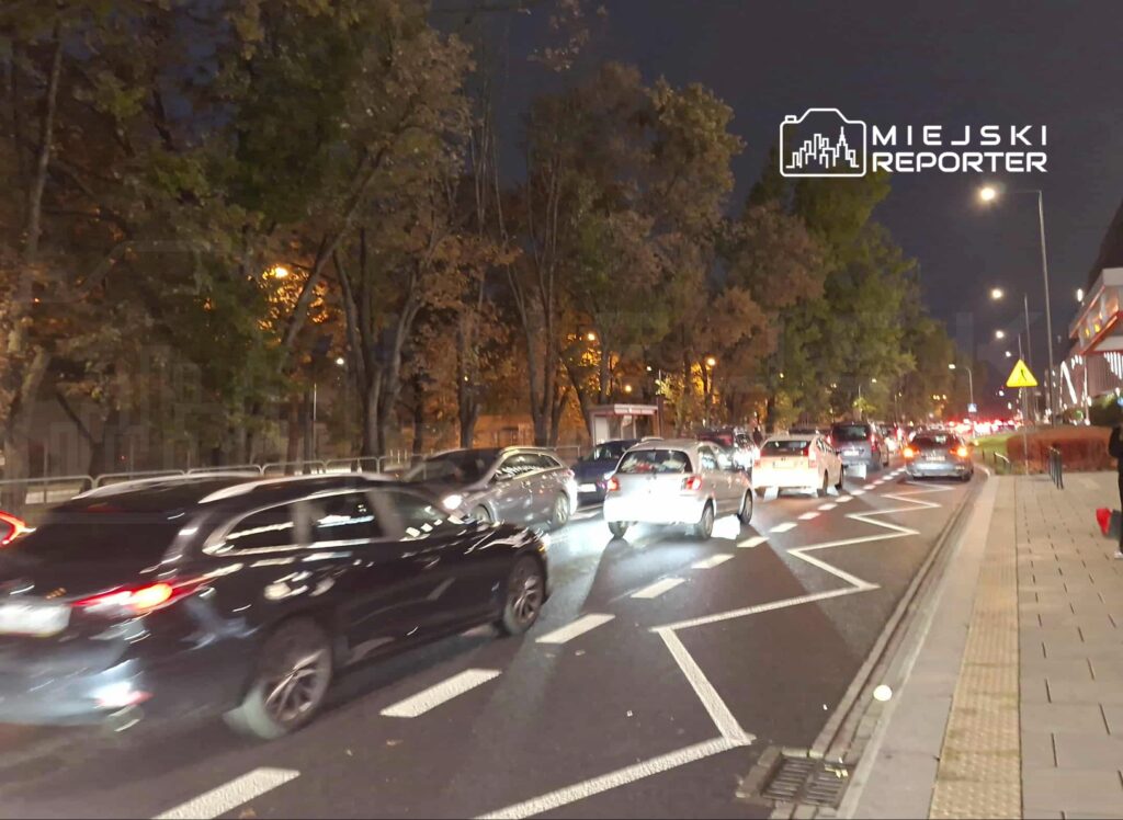 Ruch samochodowy wzdłuż oświetlonej ulicy, z samochodami stojącymi w korku w nocy, otoczone drzewami.