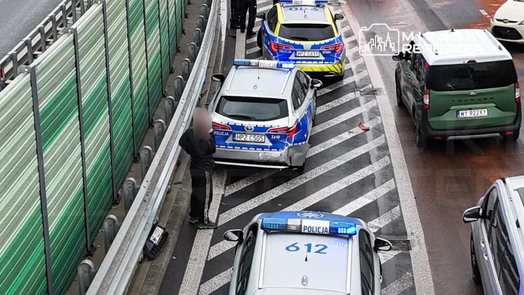 Policjant rozmawia z kierowcą przy zatrzymanym radiowozie na ruchliwej drodze. W tle widoczne inne pojazdy.