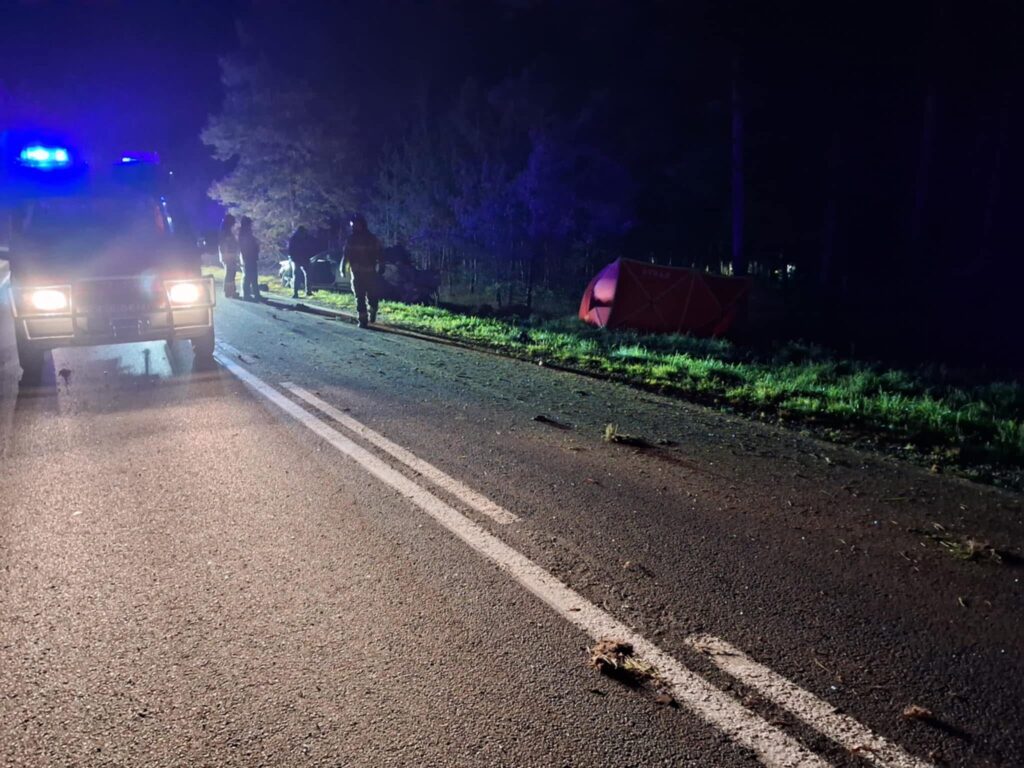 Pojazd służb ratunkowych z włączonymi sygnałami świetlnymi na drodze, obok grupy ratowników działających w nocy.