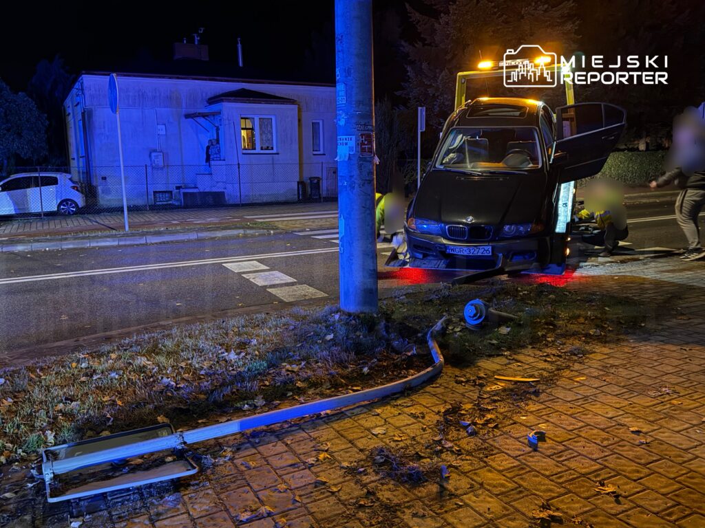 Czarny samochód osobowy stoi na poboczu drogi, a obok niego pracuje ekipa ratunkowa przy uszkodzonej latarni.