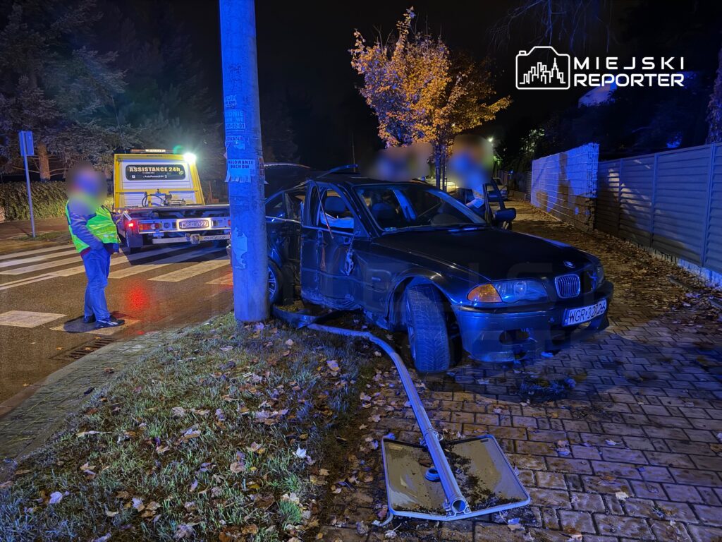 Policjant w odblaskowej kamizelce obserwuje uszkodzony samochód osobowy przy latarni, obok stoi laweta.