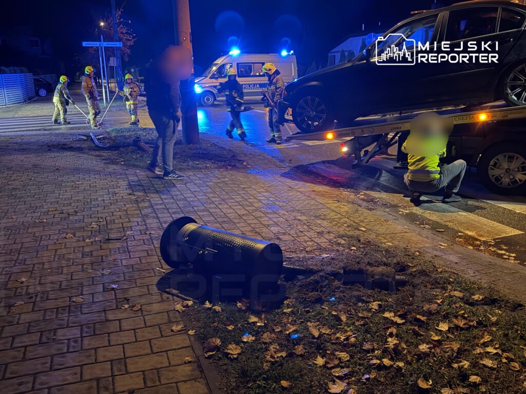 Grupa strażaków pracuje przy przewróconym koszu na śmieci, obok stoi policyjny radiowóz, a auto jest wciągane na lawetę.