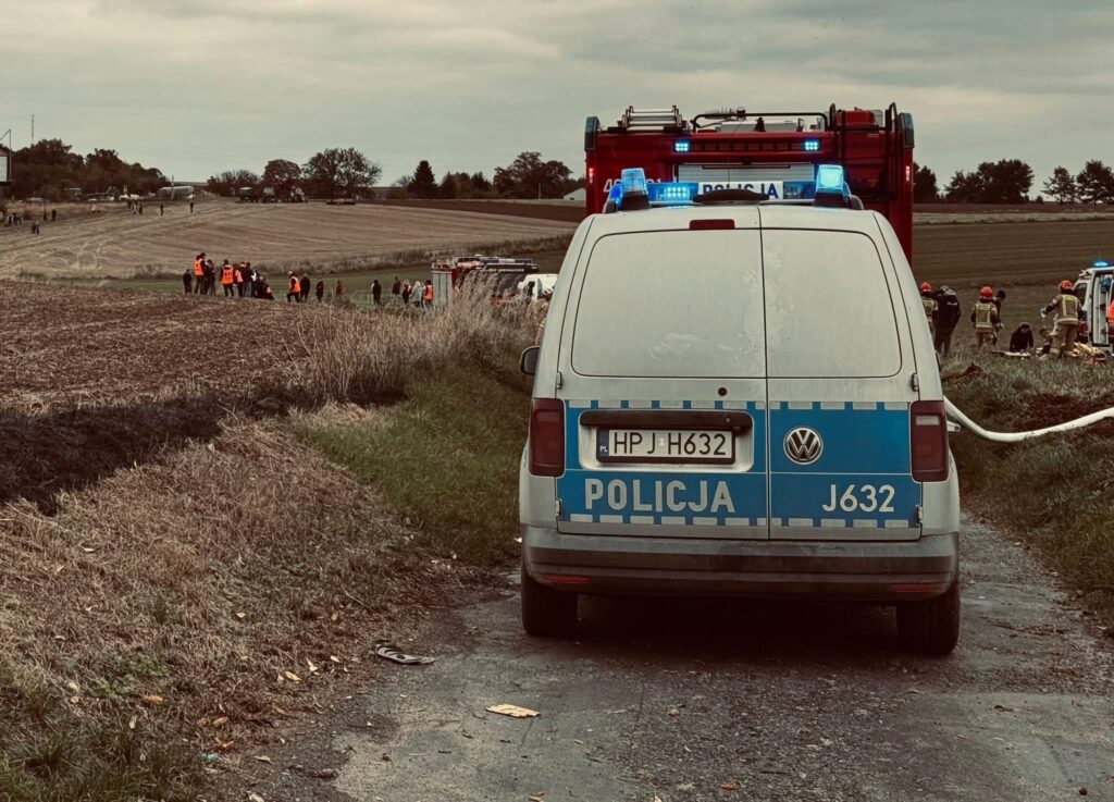 Policyjny samochód z niebieskimi światłami zaparkowany na wiejskiej drodze, w tle grupa ludzi w pomarańczowych kamizelkach.