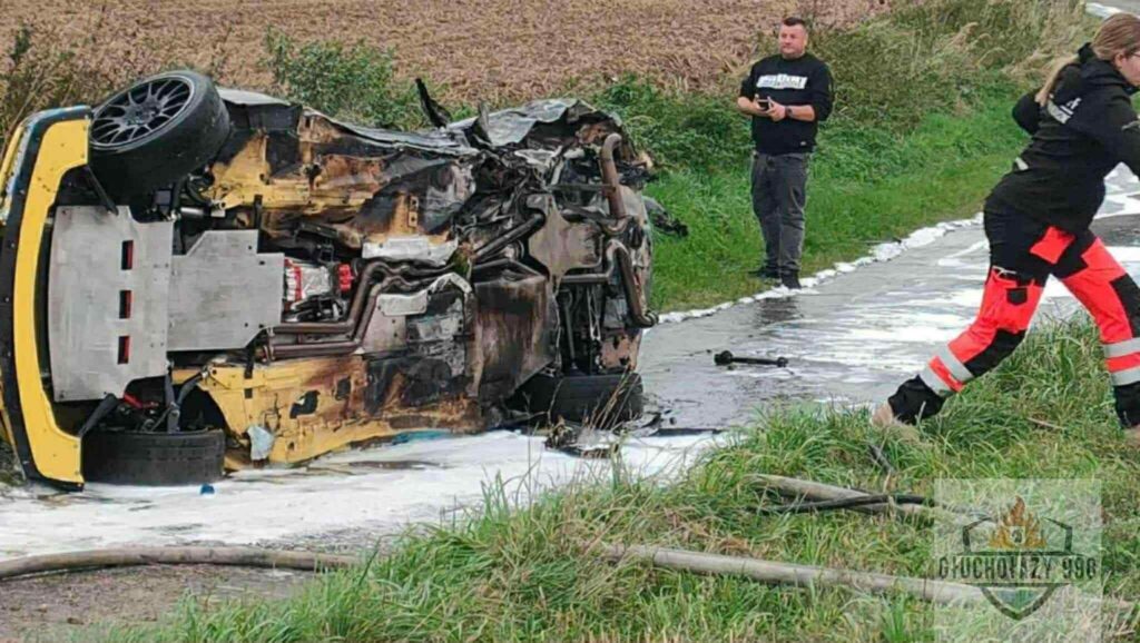Zniszczony samochód leży na boku na drodze, obok pracują strażacy w odzieży ochronnej, a w tle widać pole.