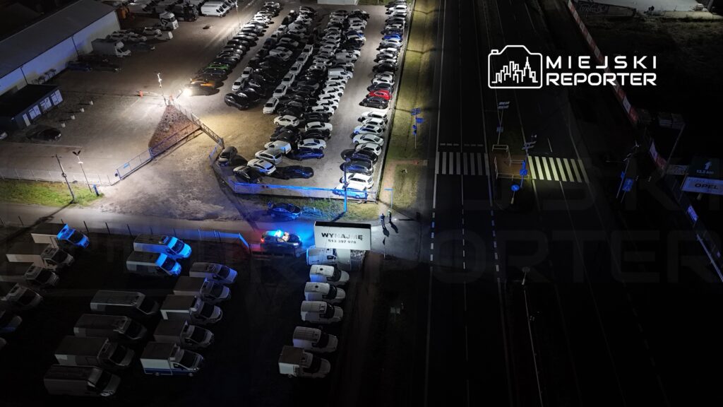 Zespół policji w niebieskich mundurach prowadzi czynności na terenie parkingu, otoczonego zaparkowanymi pojazdami.