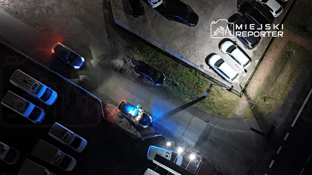 Radiowóz policyjny z włączonymi syrenami stoi na drodze, a w tle widoczne są zaparkowane samochody.