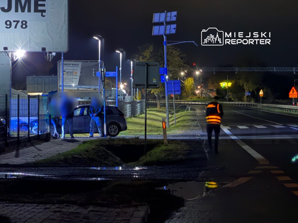 Trzech mężczyzn rozmawia obok zaparkowanego samochodu na oświetlonym poboczu drogi w nocy.