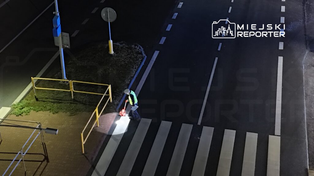 Mężczyzna w odblaskowej kamizelce mierzy długość przejścia dla pieszych przy oświetlonej ulicy.