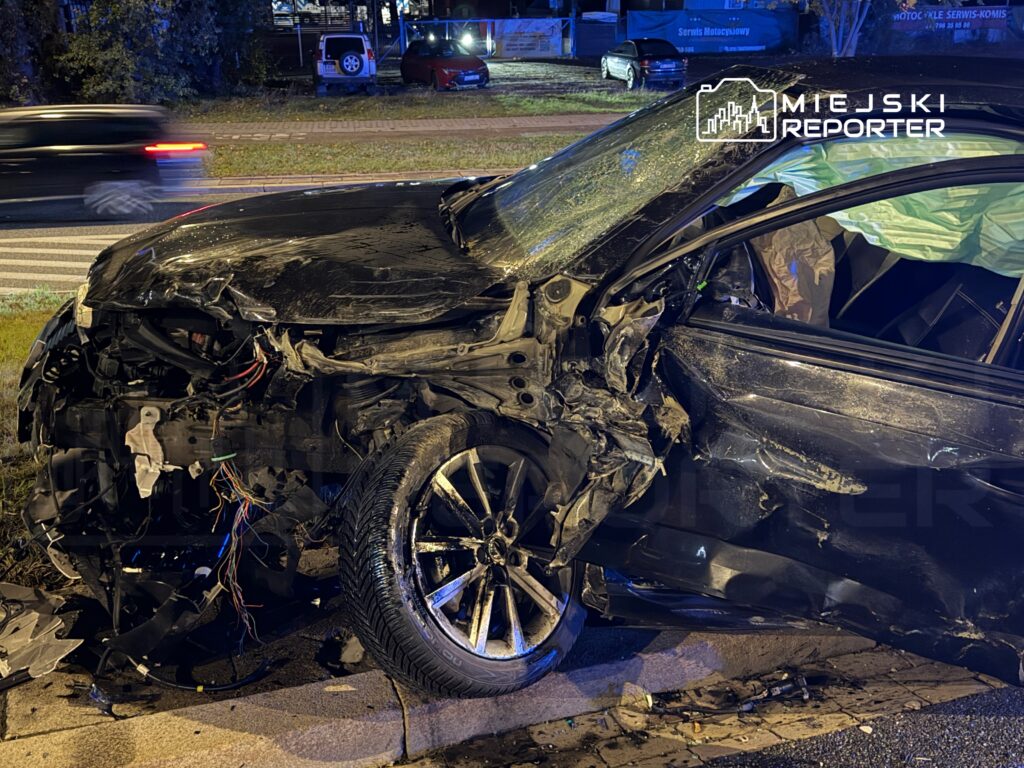 Zniszczony czarny samochód osobowy leży na poboczu drogi, z odkształconą karoserią i widocznymi przewodami elektrycznymi.