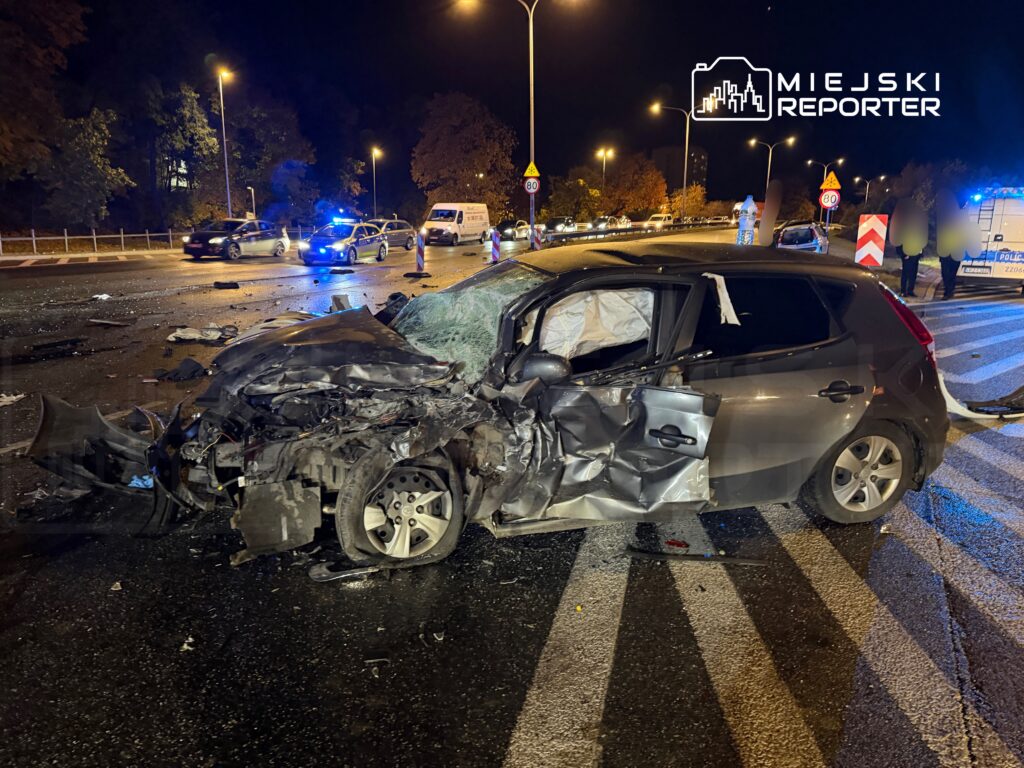 Zniszczony samochód osobowy z wygiętą karoserią leży na jezdni, otoczony przez policyjne radiowozy i oznakowanie drogi.