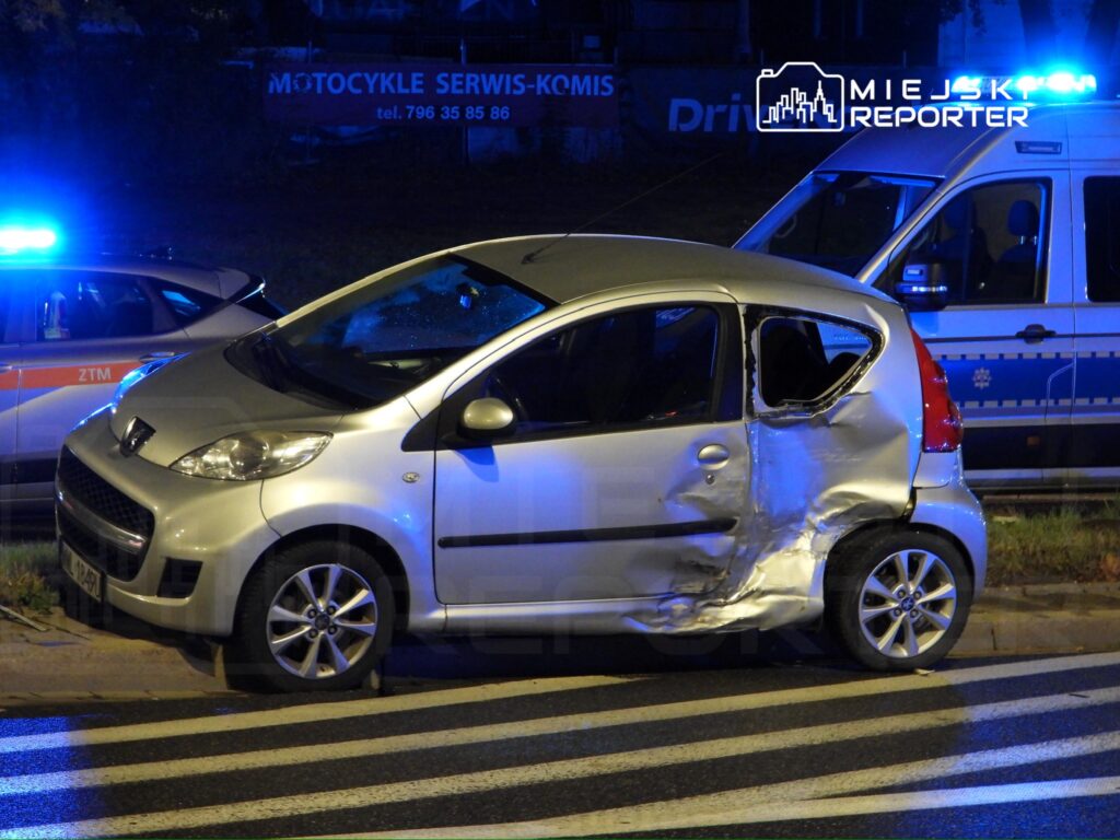 Uszkodzony srebrny samochód osobowy zaparkowany przy drodze, obok pojazdów policyjnych z włączonymi sygnałami świetlnymi.