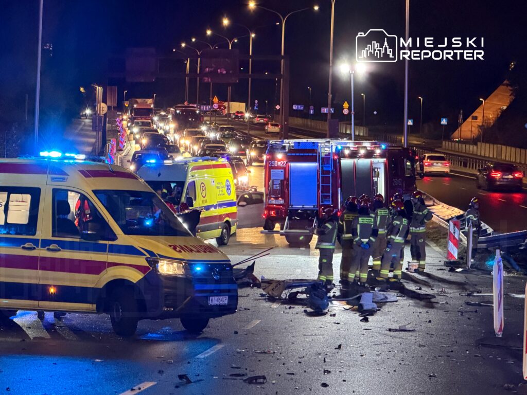 Karetka pogotowia oraz strażacy pracują na miejscu wypadku na drodze, otoczeni przez zablokowany ruch.