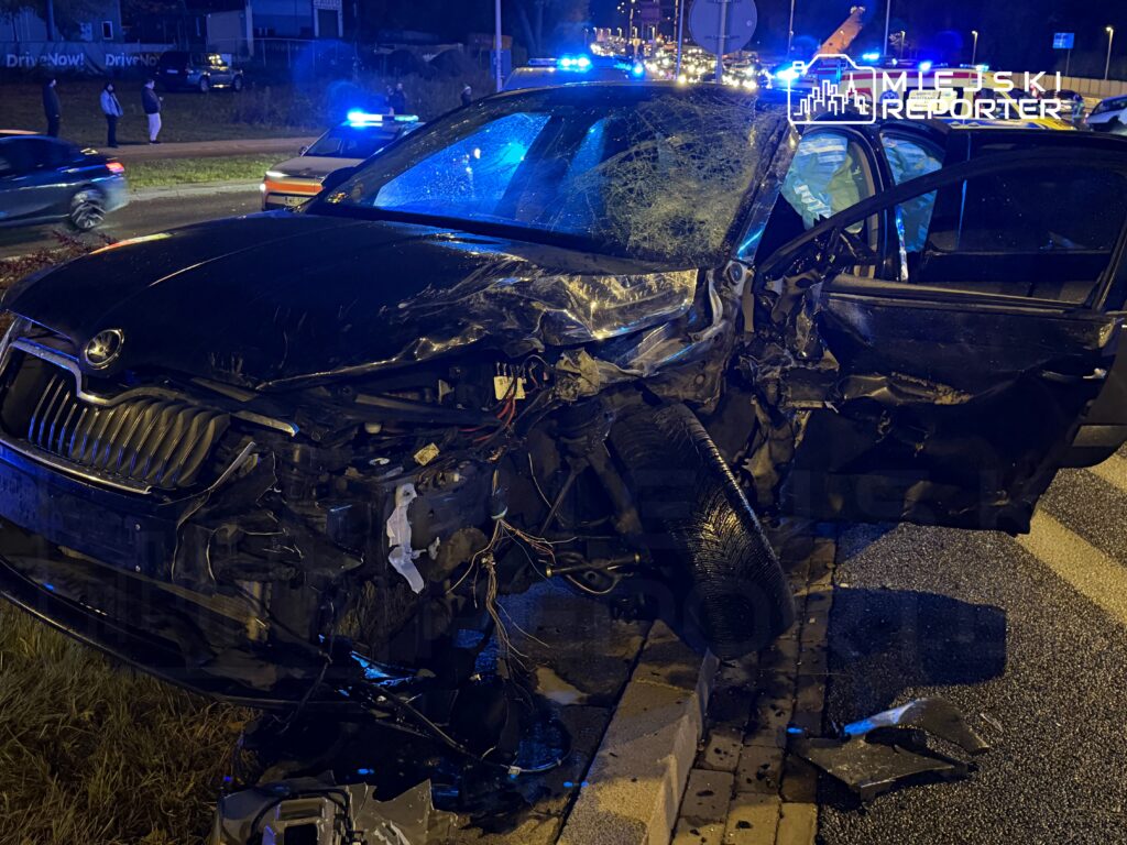 Zniszczony czarny samochód osobowy stojący na poboczu drogi, otoczony przez policyjne pojazdy z włączonymi sygnalizatorami.