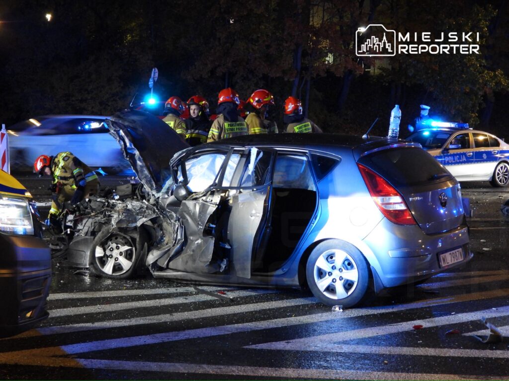 Strażacy pracują nad rozbitym samochodem osobowym na jezdni, w tle widać radiowóz policji oraz migające światła.
