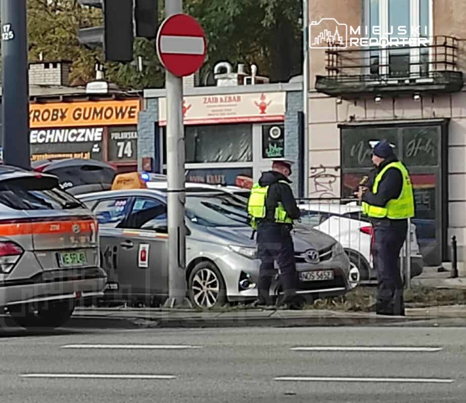 Dwaj policjanci w odblaskowych kamizelkach rozmawiają obok zaparkowanych samochodów na ulicy, w tle widoczny lokal gastronomiczny.