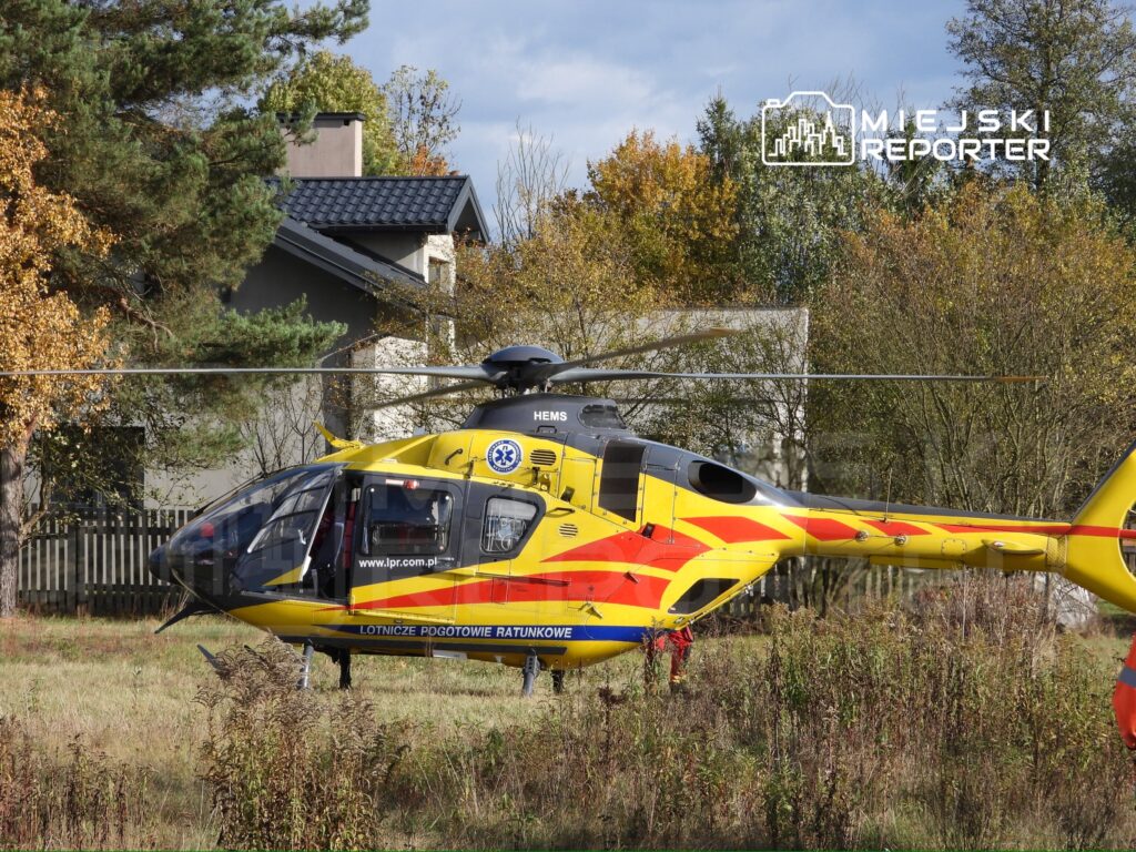 Śmigłowiec ratunkowy w kolorze żółto-czerwonym ląduje na polanie otoczonej drzewami i budynkami mieszkalnymi.