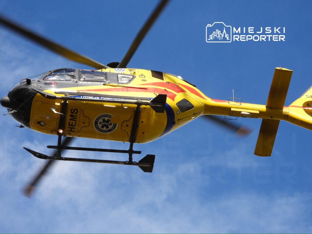 Żółty śmigłowiec ratunkowy HEMS przelatuje nad miastem, wirniki kręcą się w błękitnym niebie.
