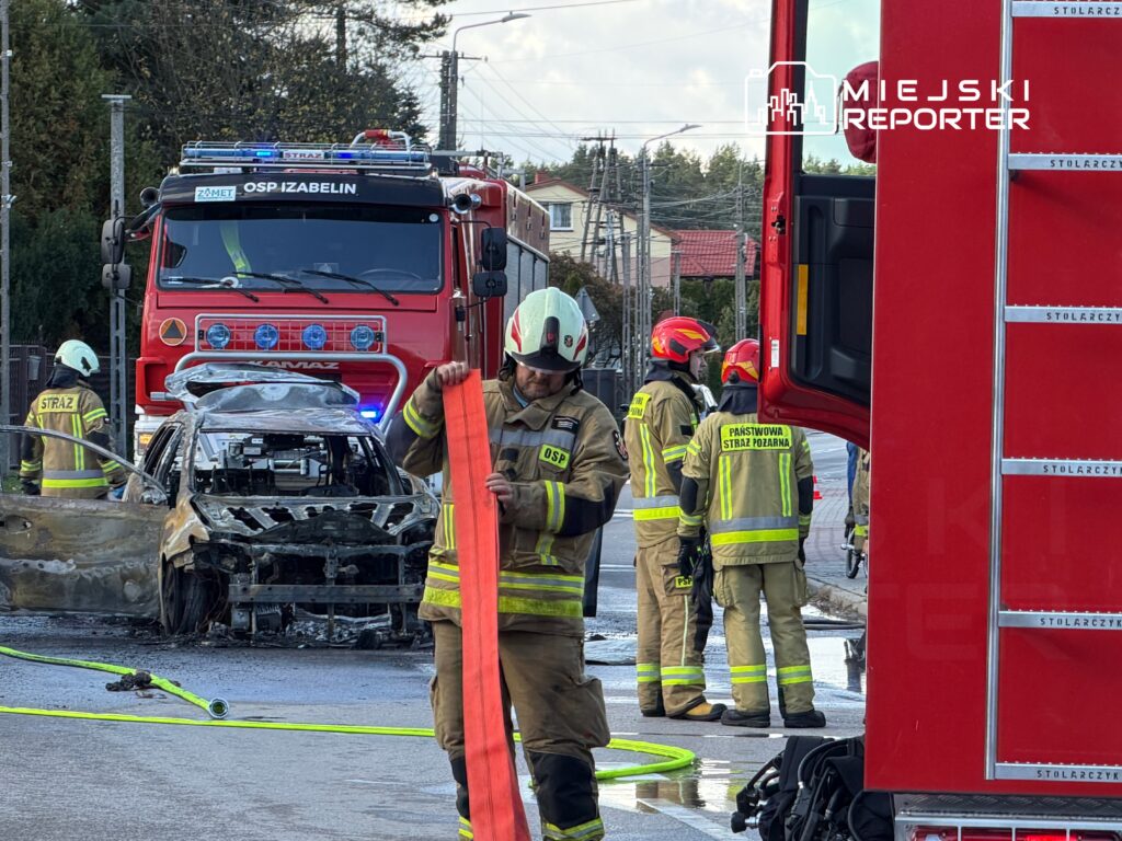 Strażacy w mundurach pracują przy spalonym samochodzie na ulicy, obok stoi wóz strażacki.