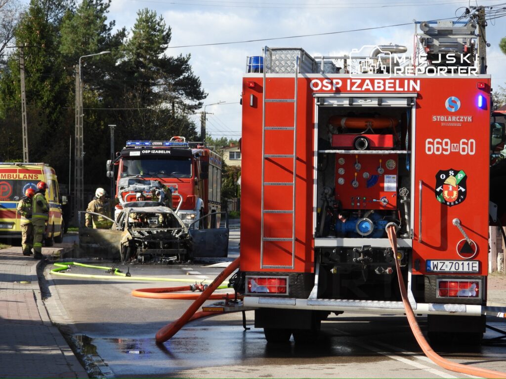Strażacy w mundurach gaszą palący się wrak samochodu na ulicy, obok stoi wóz strażacki.