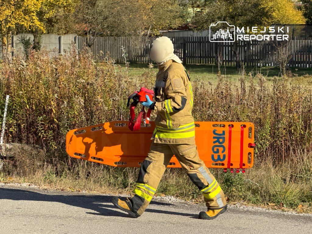 Strażak w mundurze niesie pomarańczową nosze medyczne na tle zieleni i ogrodzenia.
