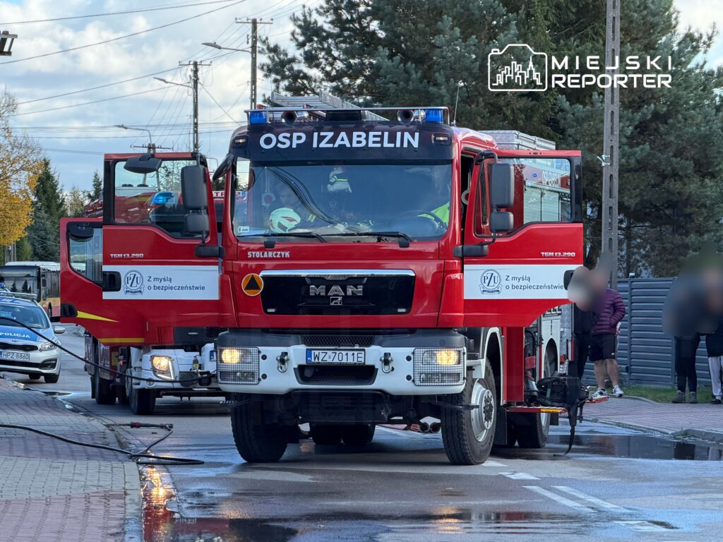 Czerwony wóz strażacki z napisem "OSP Izabelin" zatrzymany na ulicy, obok stoi radiowóz policyjny.