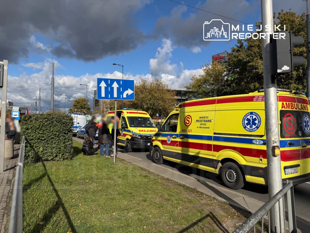 Zespół ratunkowy w żółtych ambulansach zatrzymany przy ulicy, obok grupy ludzi i znaku drogowym wskazującym kierunek.