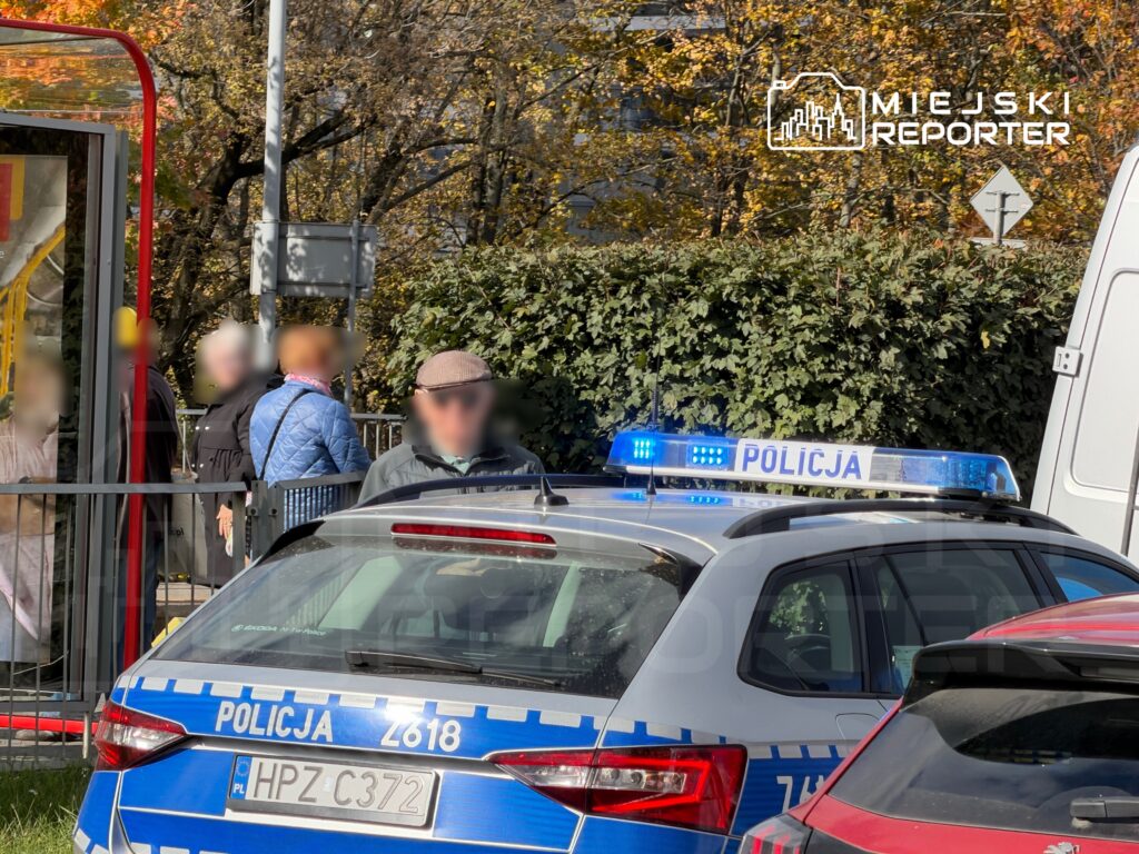 Grupa osób czeka na przystanku autobusowym obok radiowozu policji z włączonymi sygnałami świetlnymi.