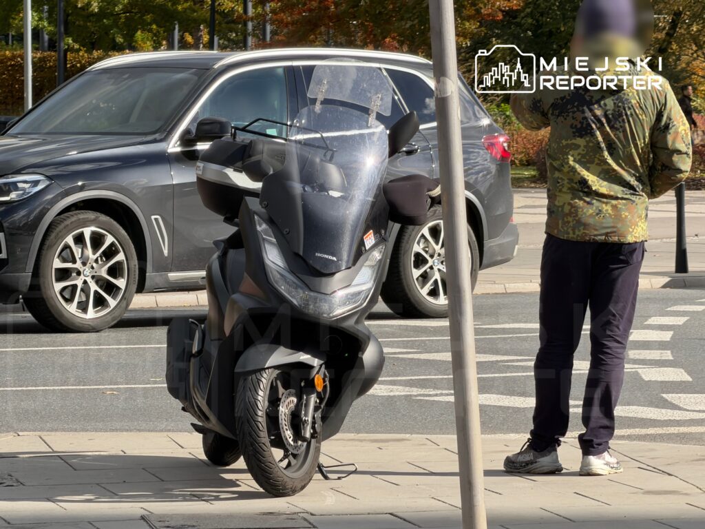 Mężczyzna w kamuflażowej kurtce rozmawia przez telefon obok zaparkowanego skutera w miejskim otoczeniu.