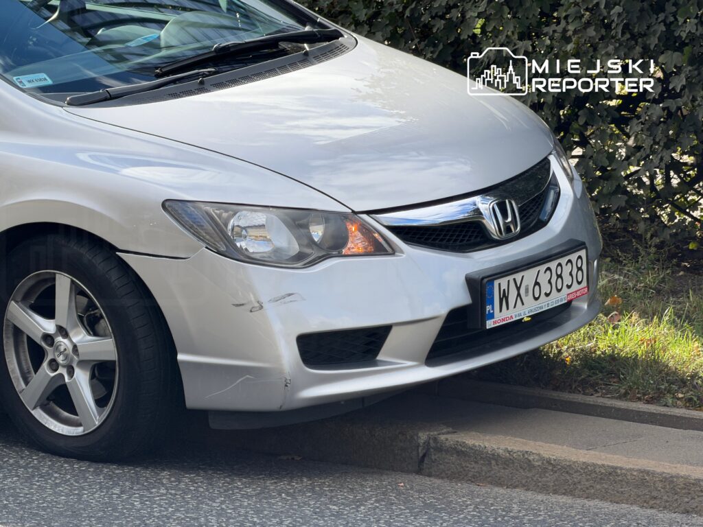 Biały samochód osobowy marki Honda zaparkowany na krawężniku, z widocznymi zadrapaniami na przednim zderzaku.