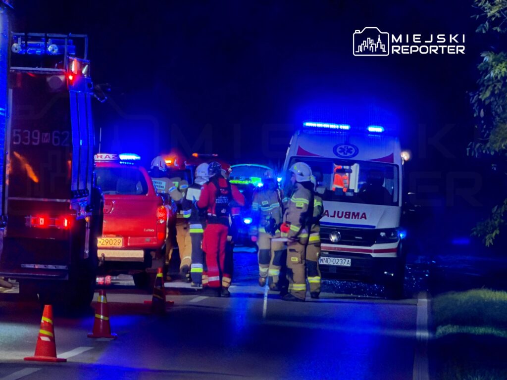 Grupa ratowników medycznych i strażaków pracuje na miejscu wypadku, otoczona pojazdami ratunkowymi z włączonymi sygnałami.