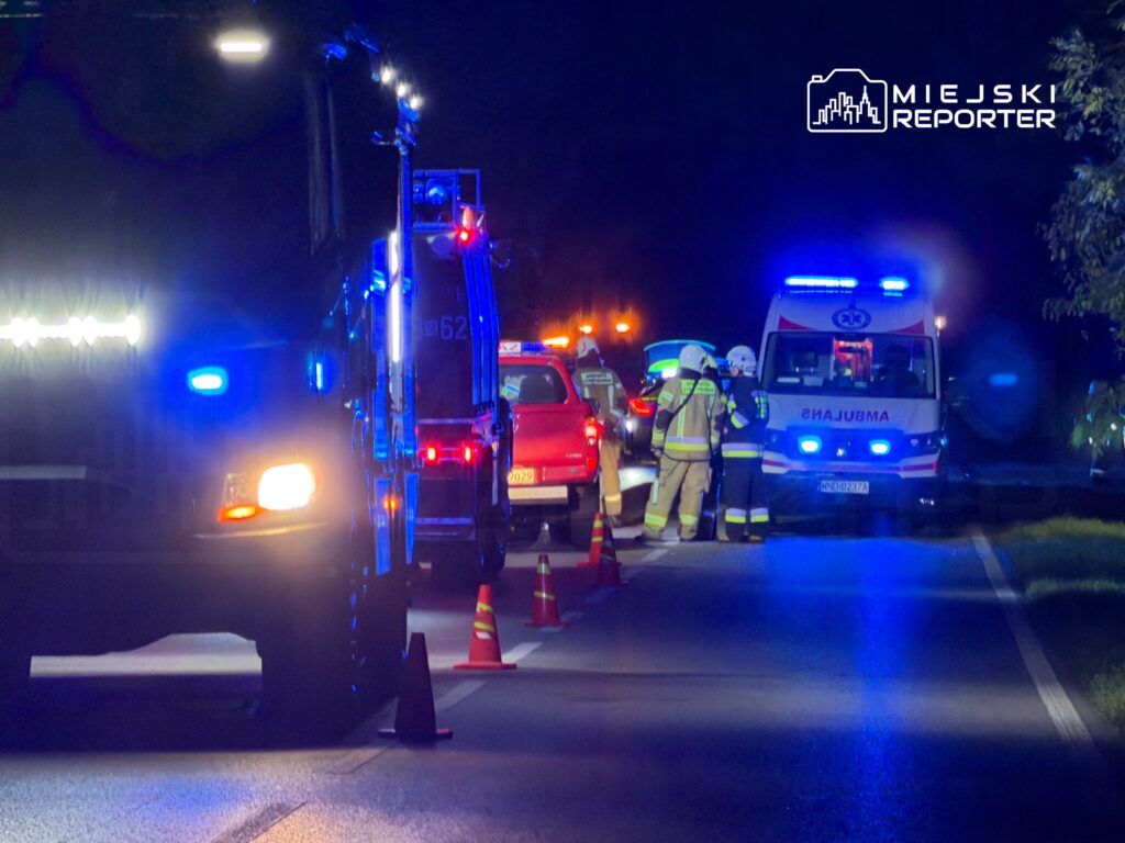 Akcja ratunkowa w nocy, z zespołem ratowników medycznych i strażaków przy wozie strażackim oraz ambulansie.