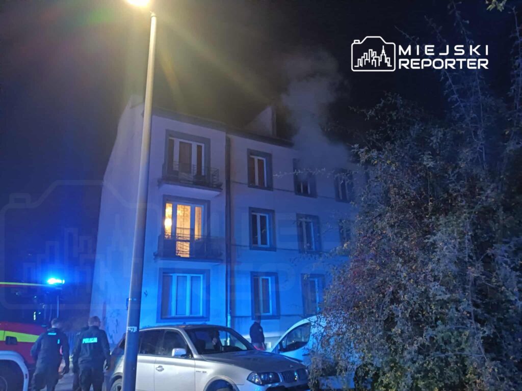 Dym wydobywający się z okna budynku mieszkalnego, w pobliżu policjanci i strażacy przy samochodach służbowych.