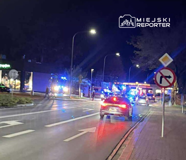 Nocna scena na ulicy z radiowozami i pojazdami straży pożarnej, oświetlonymi niebieskimi światłami.