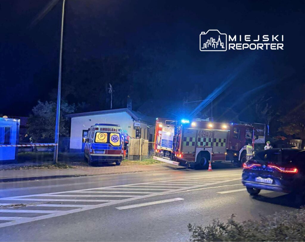 Na ulicy w nocy stoi karetka i wóz strażacki, obok grupa ratowników pracuje przy budynku.