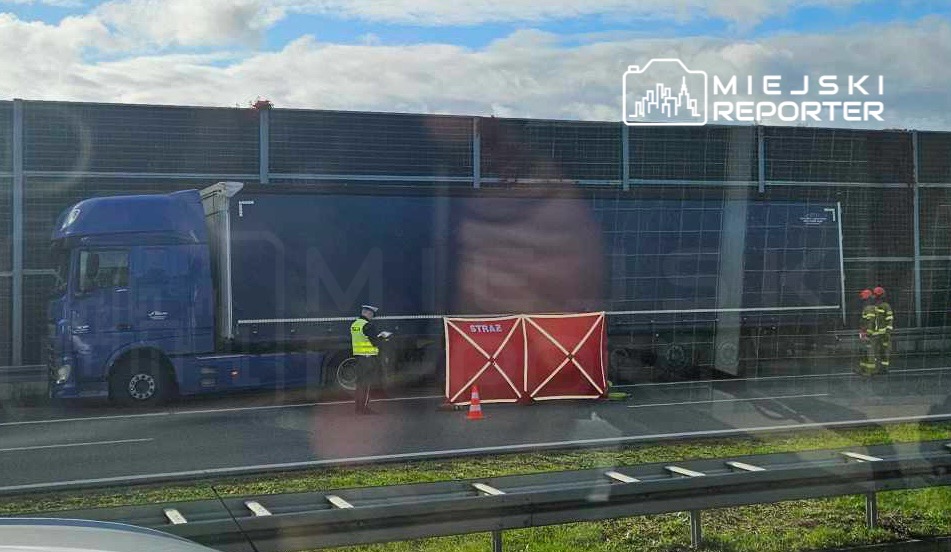 Nieoznakowany niebieski ciężarówka zatrzymana obok bariery z napisem "STRAŻ", w towarzystwie policjanta i strażaków.