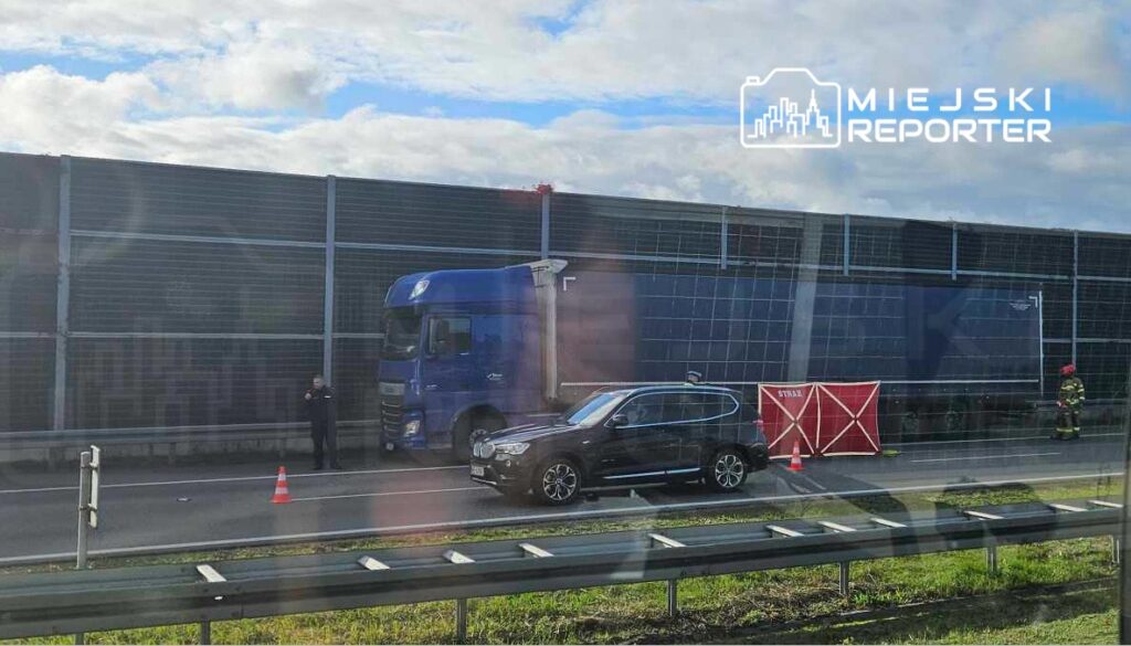 Nieoznakowany niebieski ciężarówka zatrzymana przy drodze, obok stoi policjant, a w tle widoczna czerwona bariera.