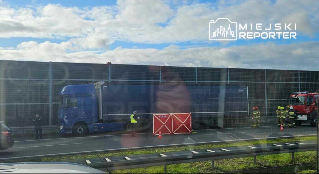 Na drodze stoi niebieska ciężarówka, obok której pracują strażacy w mundurach. Policjant kieruje ruchem, a w tle widoczna jest siatka dźwiękochłonna.