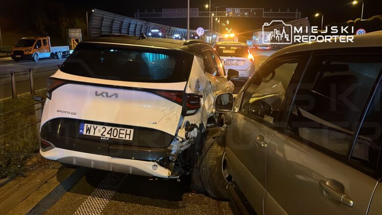 Zderzenie dwóch samochodów na pasie drogowym, jeden z pojazdów to biały Kia Sportage z uszkodzeniami tyłu.