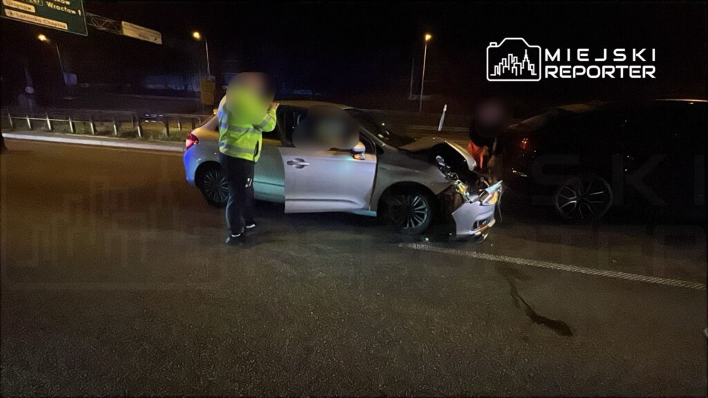 Mężczyzna w odblaskowej kamizelce rozmawia z kierowcą uszkodzonego samochodu na drodze w nocy.